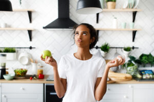 Smiled attractive mulatto girl is thinking about an apple with hilarious face and looking on the top on the white modern kitchen dressed in white t-shirt