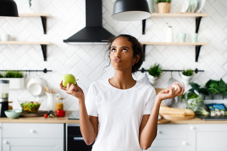 Smiled attractive mulatto girl is thinking about an apple with hilarious face and looking on the top on the white modern kitchen dressed in white t-shirt