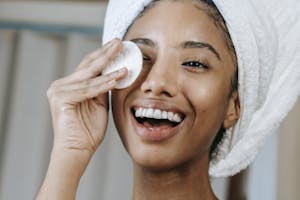 Cheerful ethnic female wrapped in towel turban moisturizing face with lotion while smiling happily | anti-aging hacks