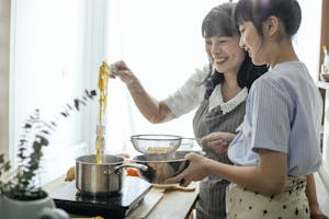 Side view of smiling Asian teenager with middle age mother serving hot boiled pasta in drainer | plant-based diet
