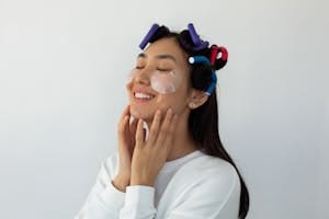 Smiling young ethnic female with hair rollers touching cheeks with eye patches on white background | double cleansing