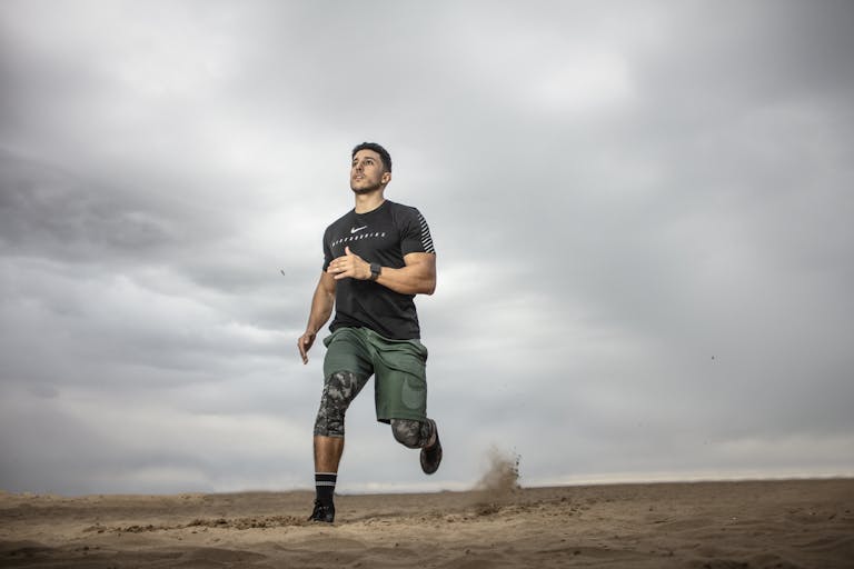 A muscular man running on sandy terrain under cloudy skies, promoting fitness and adventure. | 5 Fitness Myths Every Beginner Falls For, According to Trainers