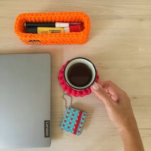 Woman's hand holding coffee on a vibrant desk with crochet container and Lenovo laptop. | Collagen Coffee