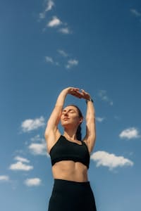 A woman in black leggings and sports bra stretches under a clear blue sky, promoting fitness and freedom.