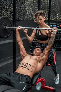 A focused young man lifting weights while a woman assists at the gym. | bench press