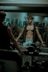 A muscular man exercises in a gym, showcasing abs in the mirror reflection, surrounded by fitness equipment. | 6 Weeks to August Abs: The EXACT Program to Carve Your Core & Get That Summer Six-Pack FAST!
