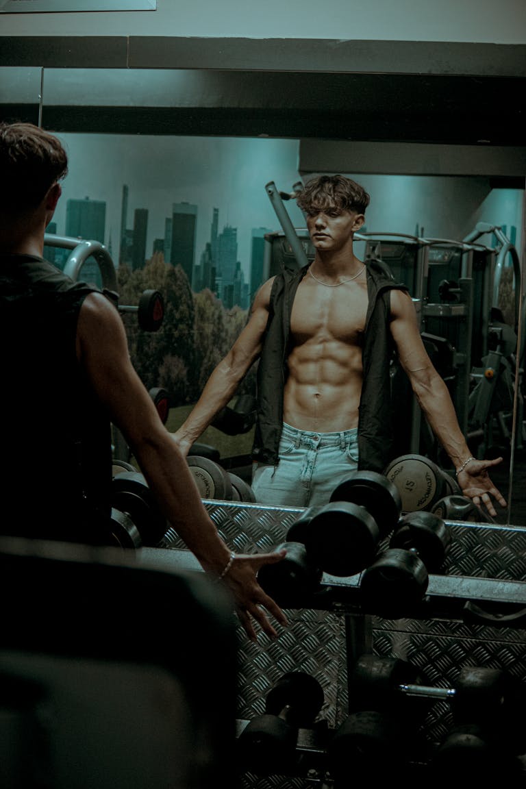 A muscular man exercises in a gym, showcasing abs in the mirror reflection, surrounded by fitness equipment.