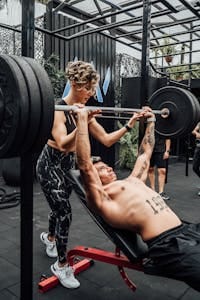 A muscular man performing a barbell bench press with assistance in a modern gym. | Can YOU Bench 225 Lbs? The True Benchmark of Strength (Are You Strong Enough?)