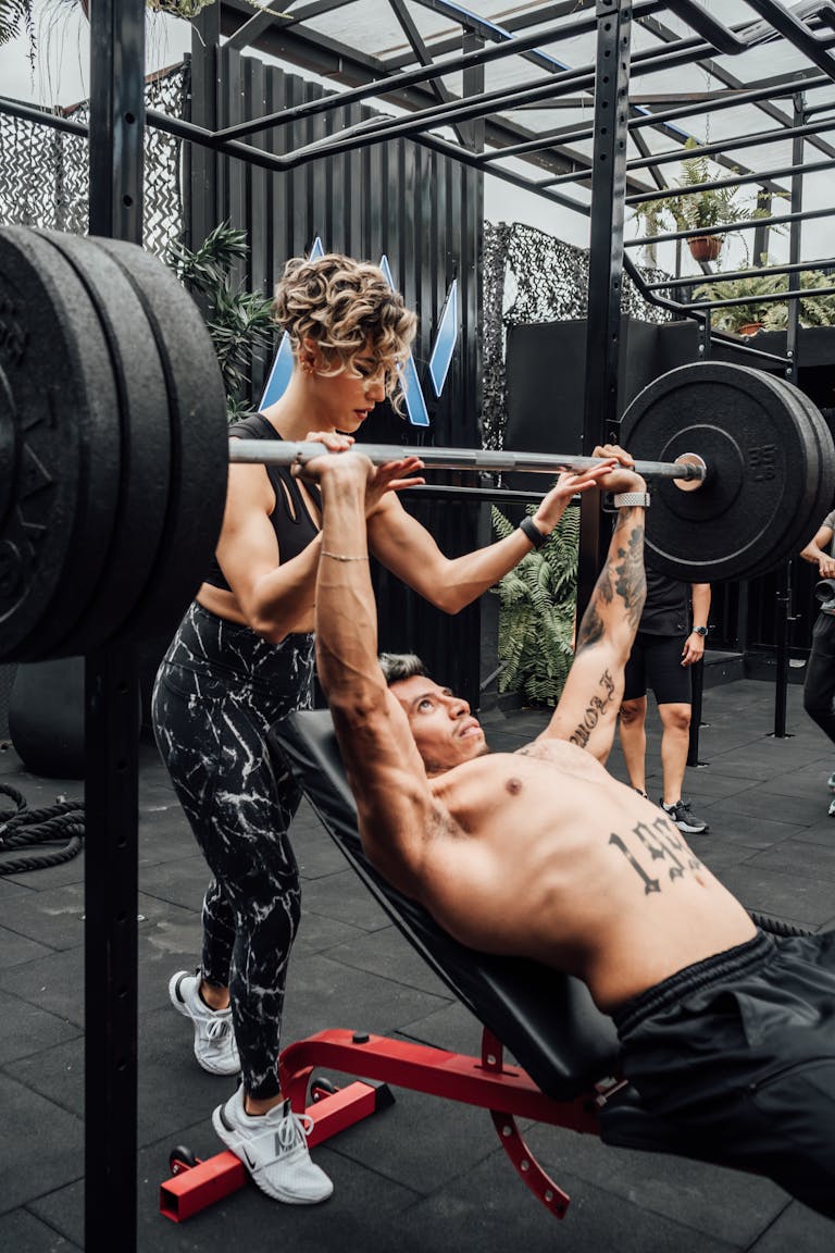 A muscular man performing a barbell bench press with assistance in a modern gym. | Can YOU Bench 225 Lbs? The True Benchmark of Strength (Are You Strong Enough?)
