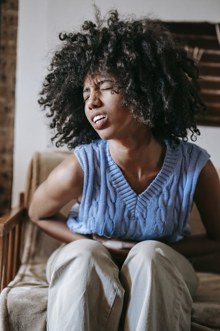 A young woman suffering from abdominal cramps, sitting indoors in discomfort. | Does Intermittent Fasting Really Cause Ulcers? Let’s Clear This Up.