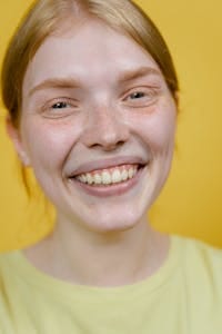 Close-up portrait of a woman smiling warmly against a vibrant yellow background. | yellow teeth