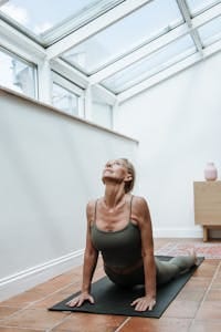 Elderly woman in yoga pose on mat in sunlit room, promoting fitness and wellness. | No Equipment, No Excuses—15 Minutes of Pilates for Full-Body Strength