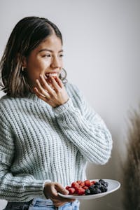 Positive Asian female in knitted sweater biting fresh healthy raspberry standing with plate of blueberries and looking away | Weight Gain vs. Berries: The Battle You Didn’t Know You Needed