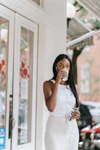 Stylish young woman sipping coffee outside a cafe, wearing earphones in an urban setting. | The Anti-Aging Supplement You Should Add to Your Coffee—Skin Benefits You’ll Love