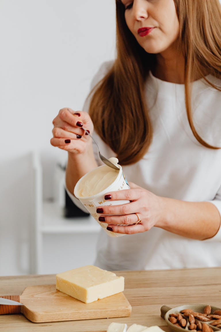 Woman with manicured nails holding yogurt container and spoon, standing by a wooden table in a kitchen. | Not All High-Protein Yogurts Are Created Equal—Here’s the Best (and the Absolute Worst)