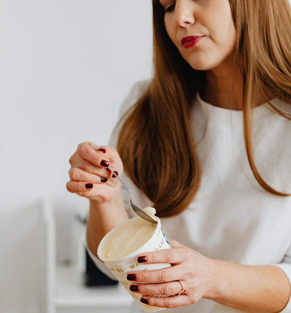 Woman with manicured nails holding yogurt container and spoon, standing by a wooden table in a kitchen. | Not All High-Protein Yogurts Are Created Equal—Here’s the Best (and the Absolute Worst)