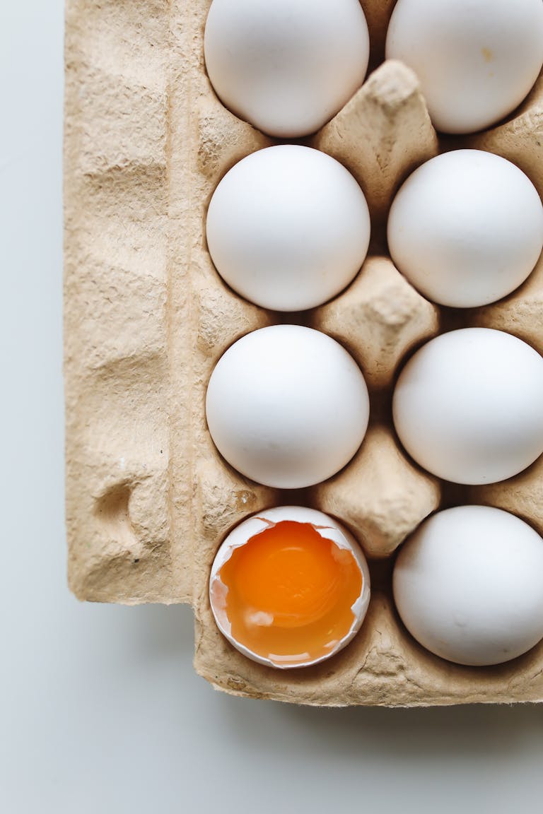 A carton of white eggs with one cracked, revealing vibrant egg yolk. Ideal for food and health themes.