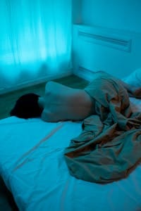 A person lies on a bed in a dimly lit, moody room, evoking solitude and tranquility. | Struggling to Sleep? This Vitamin Deficiency Could Be to Blame