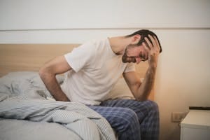 A young man in pajamas holding his head, sitting on a bed, appears to be experiencing a headache. | 10 Signs Your Body Needs More Protein