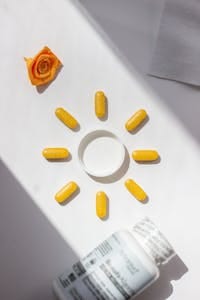 Bright yellow vitamin pills arranged in a sun shape with bottle and rose accent on white background. | Feed Your Hair: 10 Best Vitamins for Longer, Stronger Strands