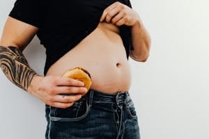 Man with tattoos holding a cheeseburger against his bare belly, wearing jeans and a black shirt. | Avoid These 6 Mistakes If You Want a Six-Pack (Stop Doing Number 2)