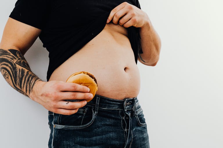 Man with tattoos holding a cheeseburger against his bare belly, wearing jeans and a black shirt. | Avoid These 6 Mistakes If You Want a Six-Pack (Stop Doing Number 2)