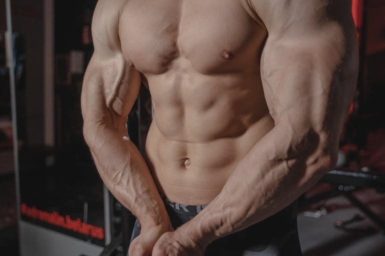 Shirtless male bodybuilder showing muscular torso and defined arms in a gym setting. | Want a Chest That Pops? Here’s the Bench Press You Should Be Doing | How To Get a Bigger Chest Without Lifting Heavy Weights