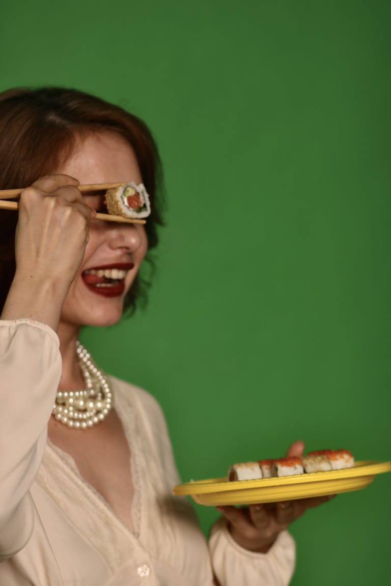 Woman holding sushi with chopsticks against a vibrant green background. | 7 Anti-Aging Foods That Experts Say You Should Eat Daily for Youthful Skin