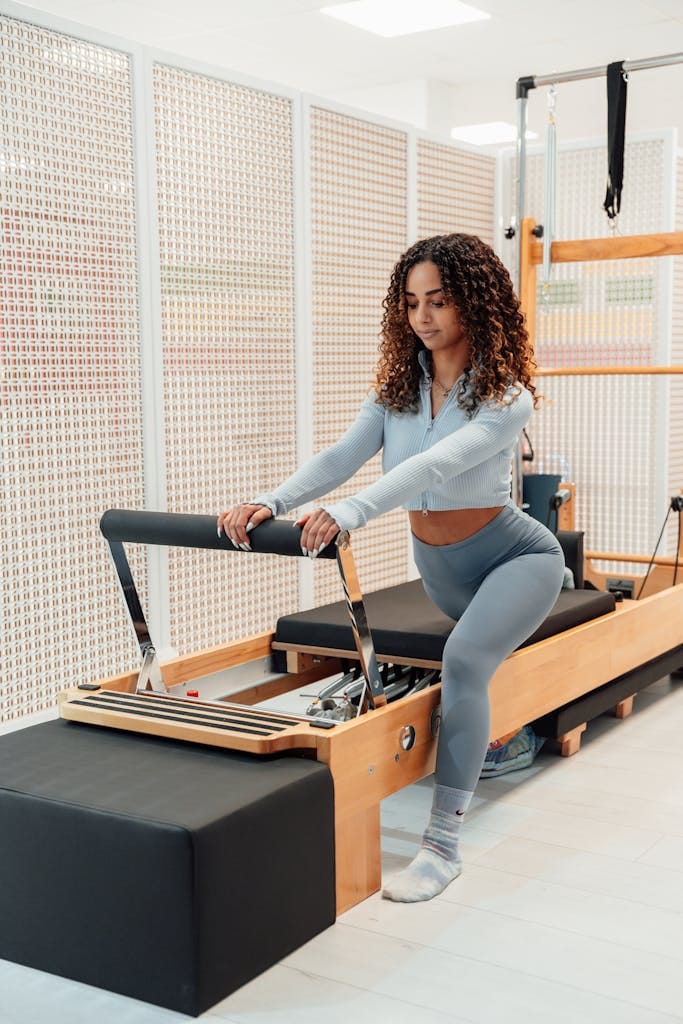 Woman with curly hair exercising on a Pilates reformer in a gym setting, wearing sportswear.