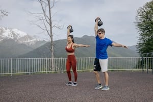 A fit couple exercises outdoors with kettlebells against a scenic mountain backdrop, showcasing a healthy lifestyle. | Skip the Gym: 4 Kettlebell Exercises That Build Serious Strength at Home