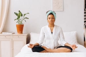 A woman meditating indoors on a bed, creating a tranquil atmosphere with a potted plant nearby. | How I De-Stress and Reconnect with My Body in 5 Minutes When Life Gets Overwhelming