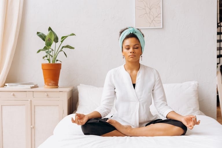 A woman meditating indoors on a bed, creating a tranquil atmosphere with a potted plant nearby. | How I De-Stress and Reconnect with My Body in 5 Minutes When Life Gets Overwhelming