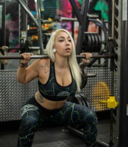 Blonde woman with tattoos working out in a gym using weightlifting equipment. | Heel-Elevated Squats