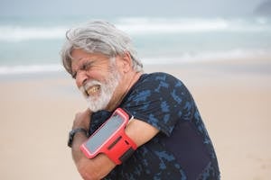 Elderly man grimacing from shoulder pain during seaside walk, showcasing health issue outdoors. | This Posture Hack Fixes Shoulder Tension in Just 60 Seconds, According to a Physical Therapist