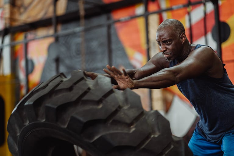 Senior man focused on pushing a large tire in an urban gym setting, highlighting fitness and strength.