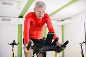 Senior man in red activewear exercising on stationary bike indoors, promoting healthy lifestyle. | Over 50? Flatten Your Belly in Just 7 Days—Trainer Shares the Secret