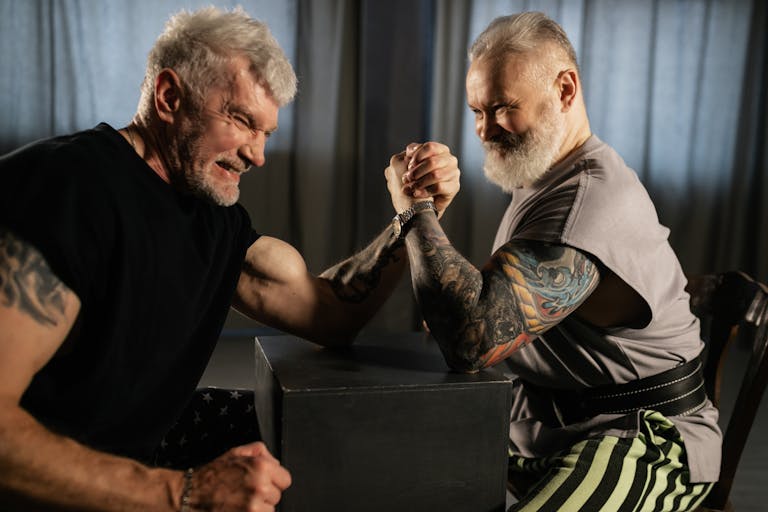 Two senior men engaged in an intense arm wrestling match indoors. | Never Too Late! A Trainer’s Top Moves to Boost Strength and Mobility After 50