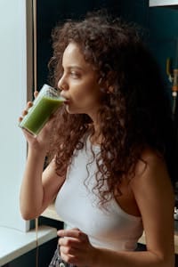 Young woman with curly hair drinking a nutritious green smoothie near a window. | Constipation Cure? This One Drink Guarantees You'll Poop Before Breakfast (Dietitian Says!)