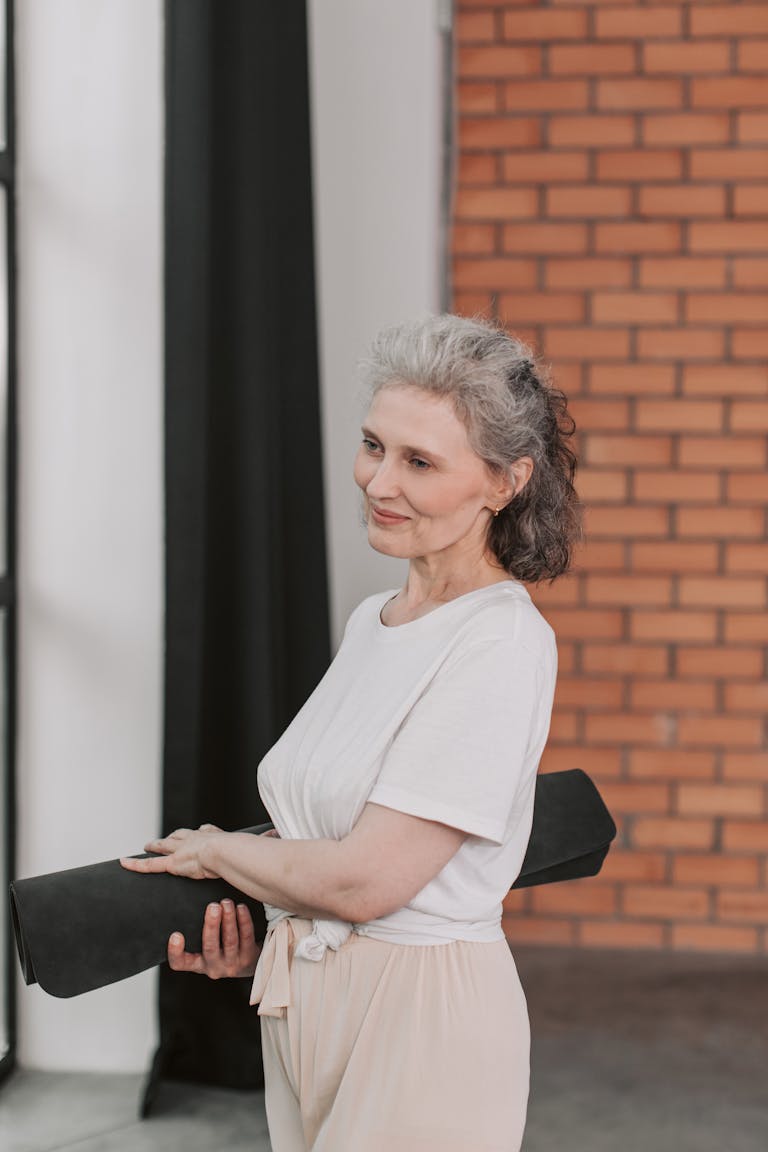 A graceful senior woman with gray hair holding a yoga mat, standing in a bright room. | Experts Say These 8 Habits Are the Real Signs You’re Aging Gracefully | Menopause Weight Gain? This 5-Move Routine Shrinks Belly Fat and Builds Muscle (Backed by Science)