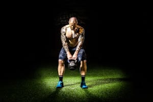 Muscular man with tattoos lifting a kettlebell indoors under a spotlight. Intense fitness and strength training. | The Kettlebell Secret That’s Making Fast Runners Even Faster