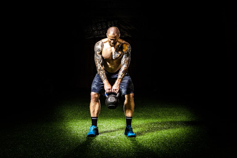 Muscular man with tattoos lifting a kettlebell indoors under a spotlight. Intense fitness and strength training. | The Kettlebell Secret That’s Making Fast Runners Even Faster