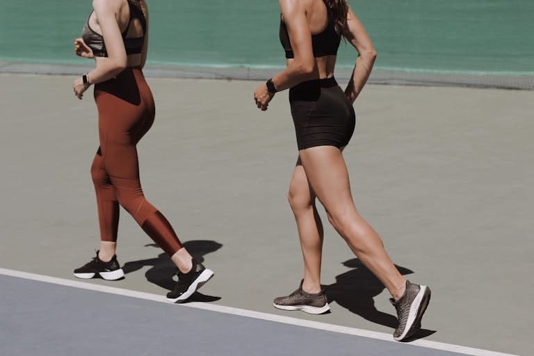 Two women walking on an outdoor track in sportswear, emphasizing healthy lifestyle and fitness. | This 7-Day Walking Plan Burns Fat Fast—And It’s Trainer-Approved