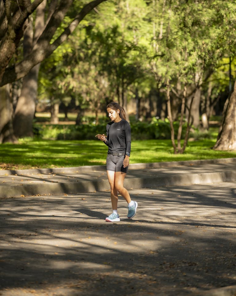 Woman jogging in a sunlit park, checking smartphone while exercising outdoors. | Struggling to Hit 10,000 Steps Again? Here’s Exactly What Worked for Me | The Desk Job Antidote: Experts Reveal How Much Exercise Truly Erases A Full Day of Sitting