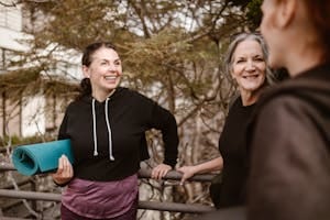 Women smiling and conversing outdoors, holding a yoga mat, embracing health and wellness.
