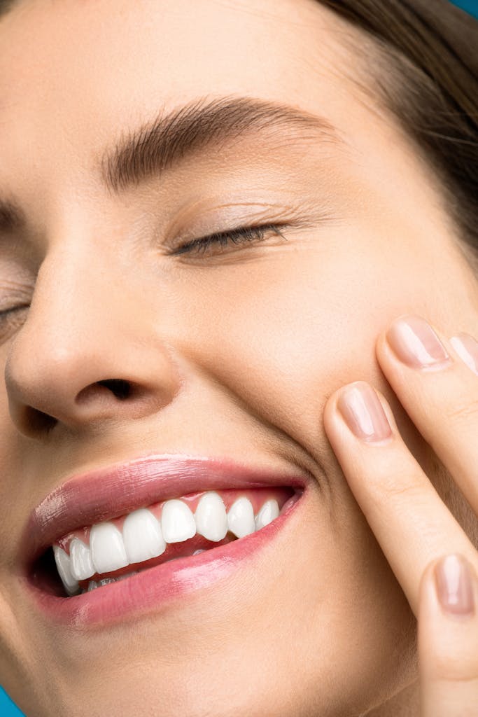 A close-up portrait of a joyful woman with closed eyes, pink lipstick, and her hand touching her cheek. | I Avoided the Dentist & Got a Celebrity Smile Using These Peroxide-Free Whitening Strips