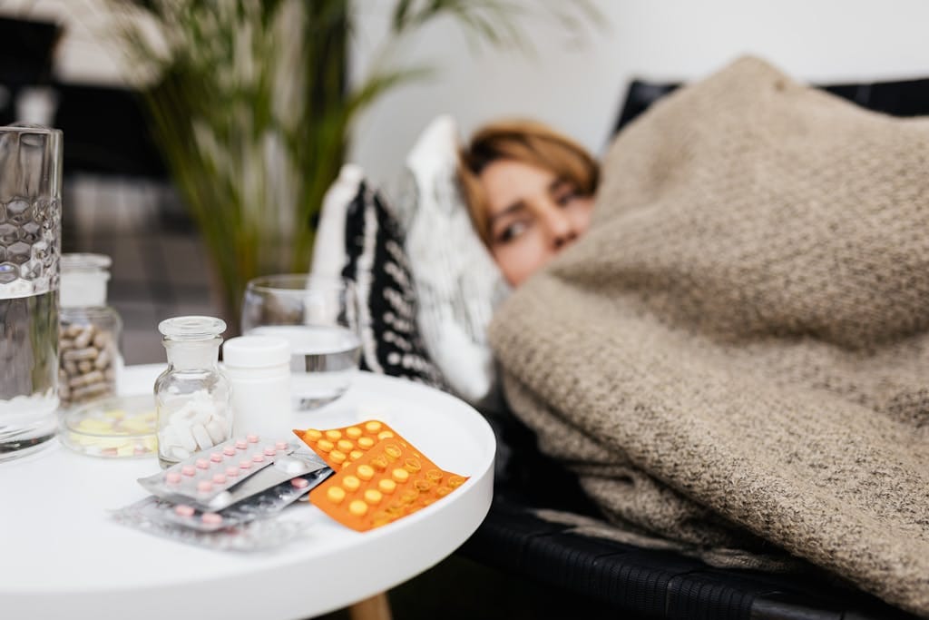 A woman rests under a blanket surrounded by medications on a table, depicting illness and recovery. | This Medication and Magnesium Don’t Mix—Here’s Why It Can Be Dangerous