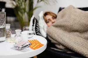 A woman rests under a blanket surrounded by medications on a table, depicting illness and recovery. | This Medication and Magnesium Don’t Mix—Here’s Why It Can Be Dangerous