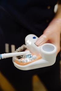 Crop unrecognizable woman wearing uniform in latex gloves using electric brush to apply toothpaste on jaw mockup for everyday teeth treatment procedure | Dentists Reveal: Is an Electric Toothbrush Really Better—Or Just Hype?