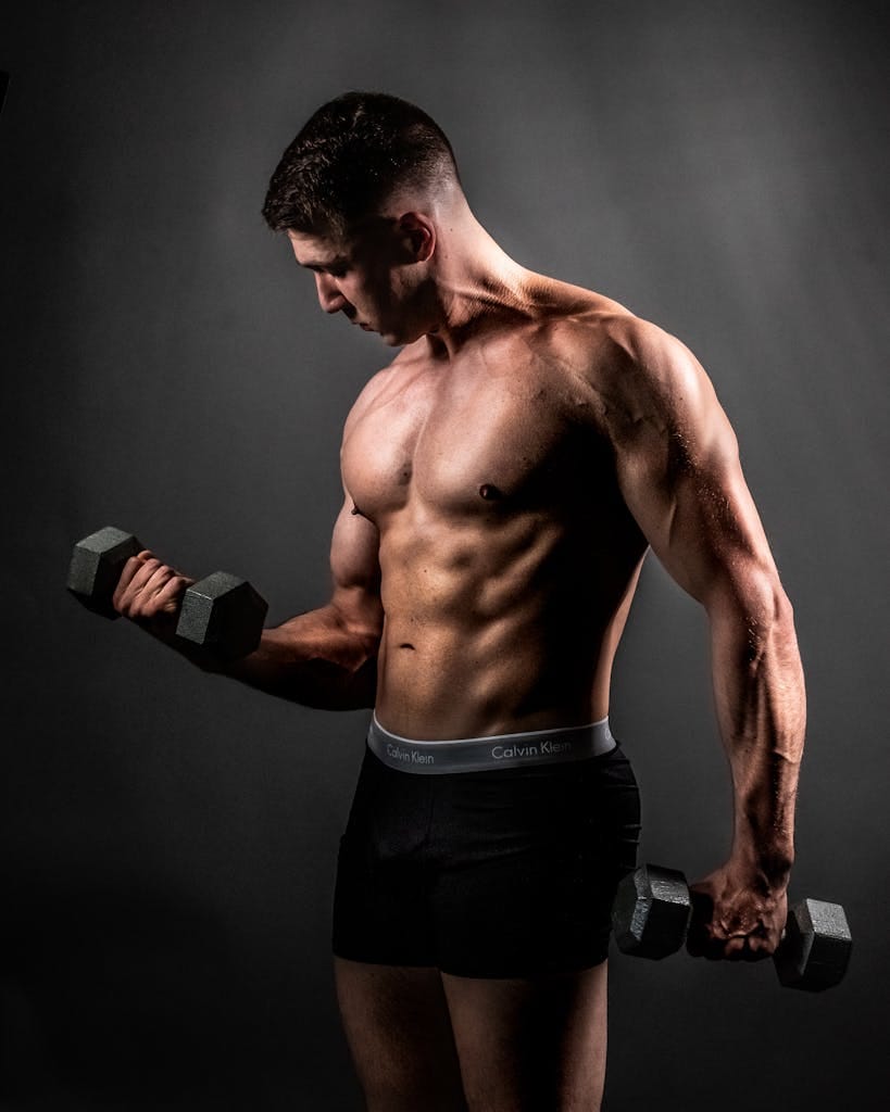 Fit shirtless man lifting dumbbells in a studio, showcasing his muscles and fitness. | Strength Training Timing: How Long Before You See Real Gains?