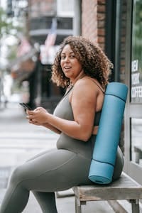 Smiling woman sitting outdoors with yoga mat, checking phone in sports attire. | No More Excuses: 7 Low-Impact Workouts Every Plus-Size Woman Can Do (Feel Strong & Confident Now!)